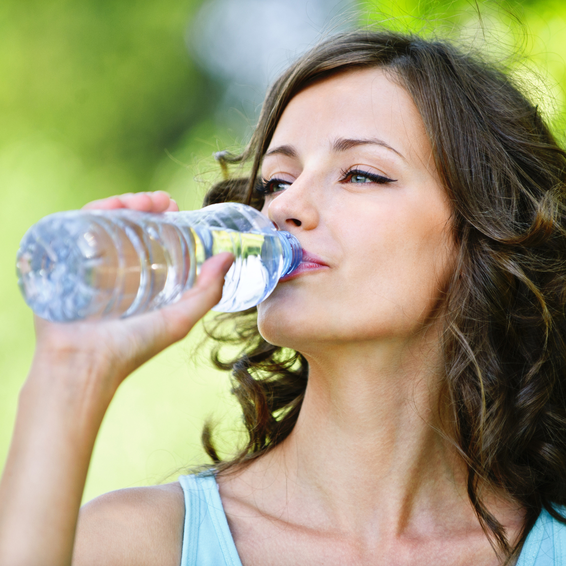 Portrait of young beautiful dark-haired woman wearing blue t-shirt drinking water at summer green park.